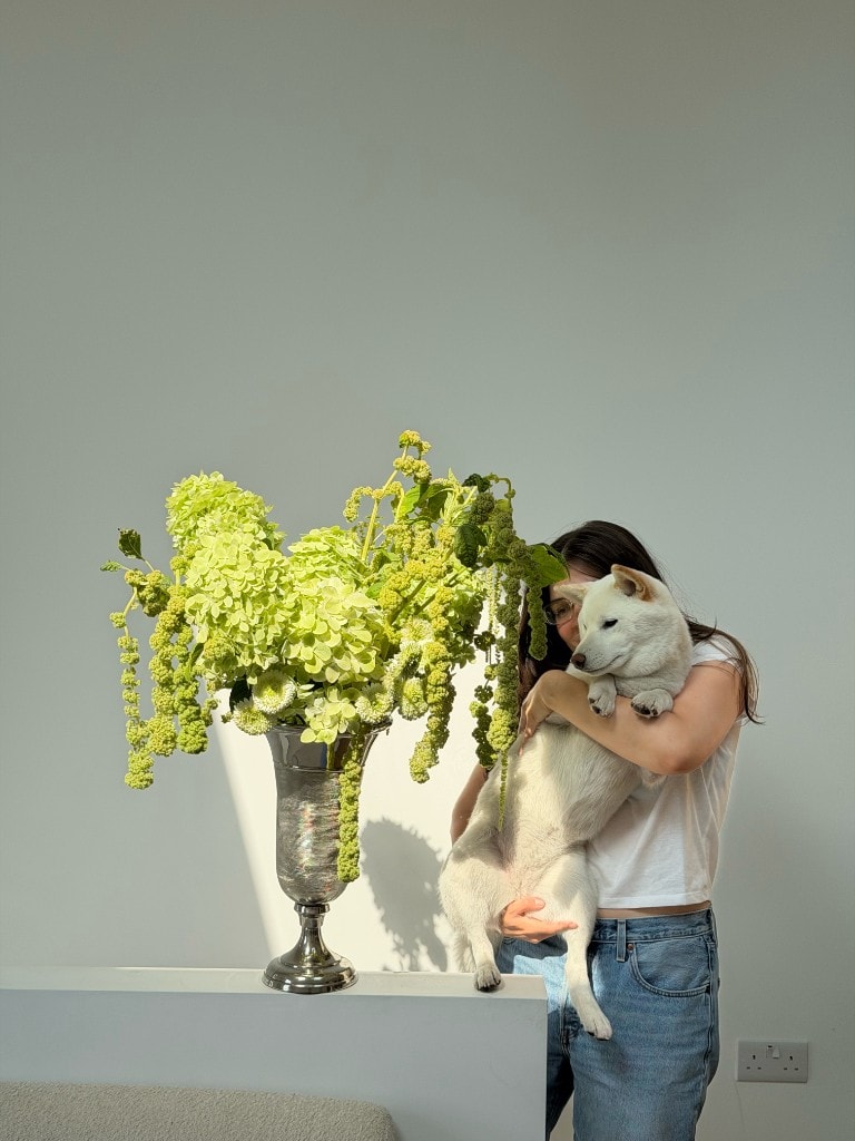 Aina holding Misha, her white studio dog, beside a sculptural green floral arrangement
