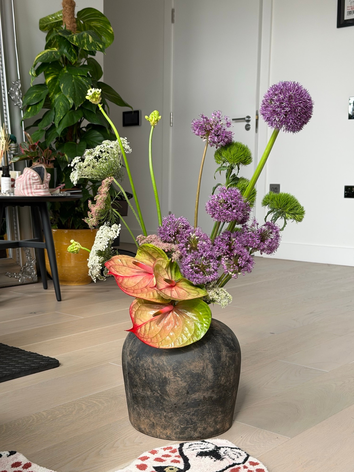 Tall sculptural arrangement of purple alliums and variegated anthuriums in a dark ceramic vase