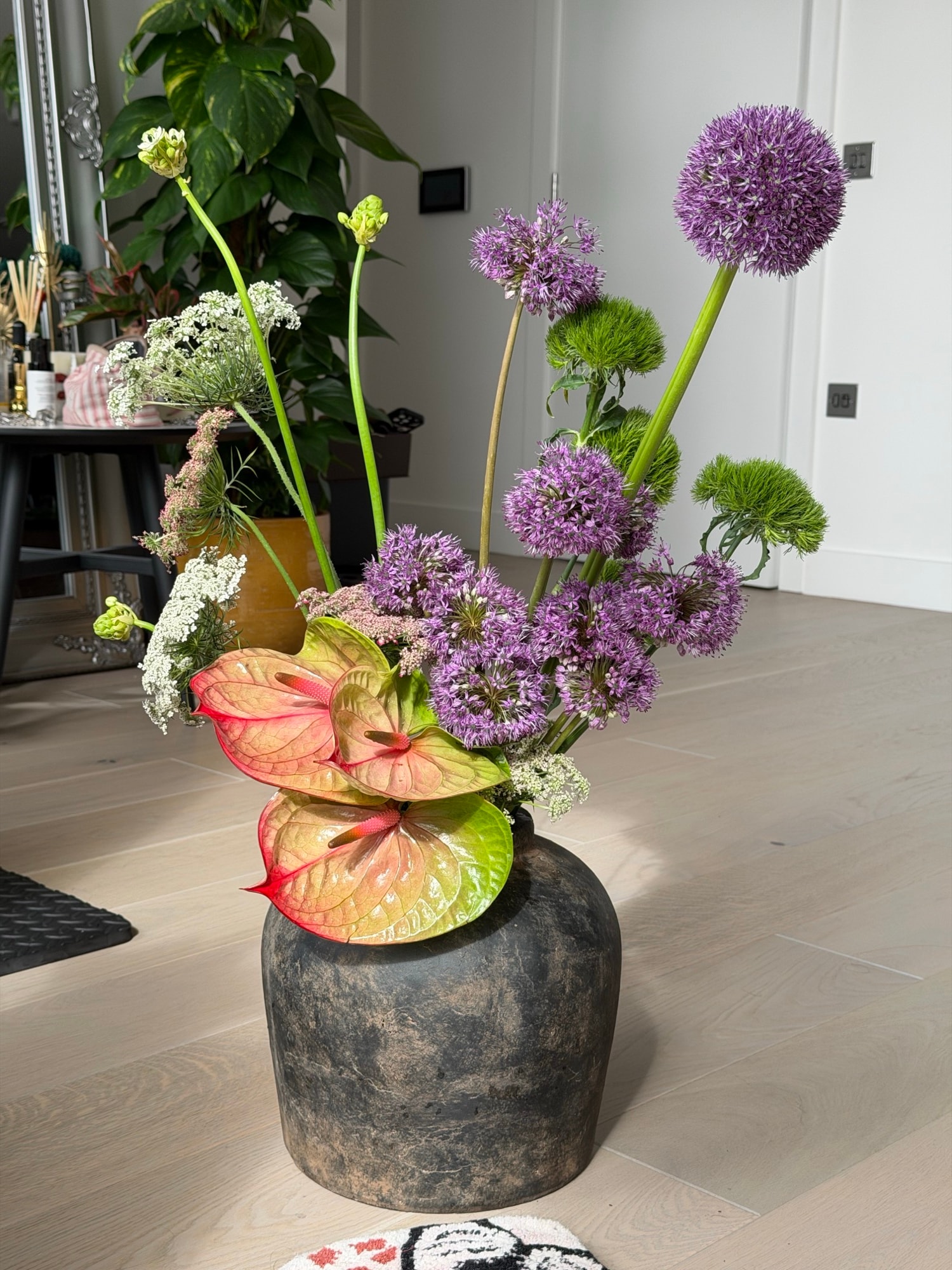 Airy allium and peach-edged anthurium arrangement in a dark rounded vase on pale wood