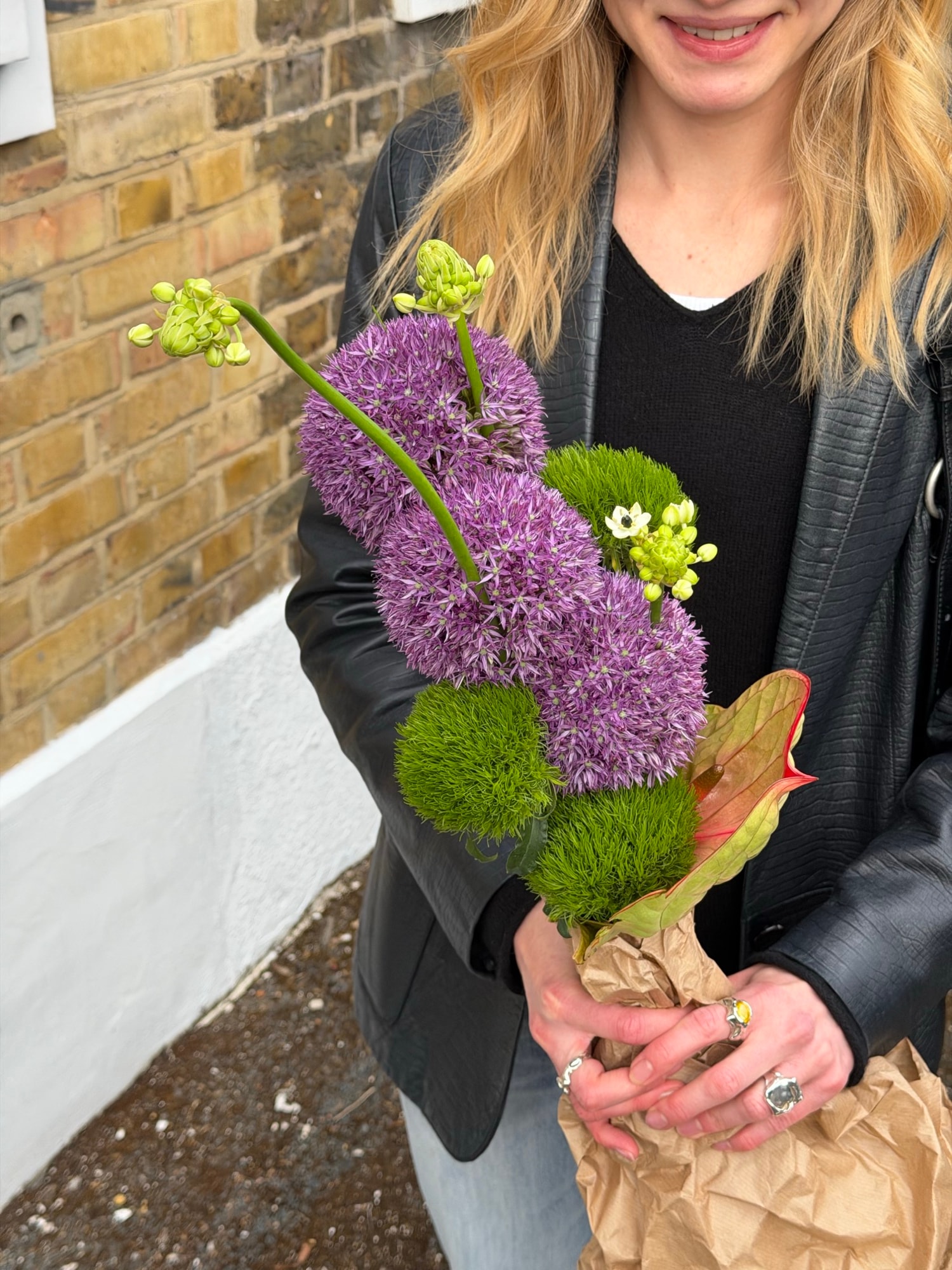 Blonde woman in a leather jacket holding a kraft-wrapped bouquet of purple alliums and green dianthus