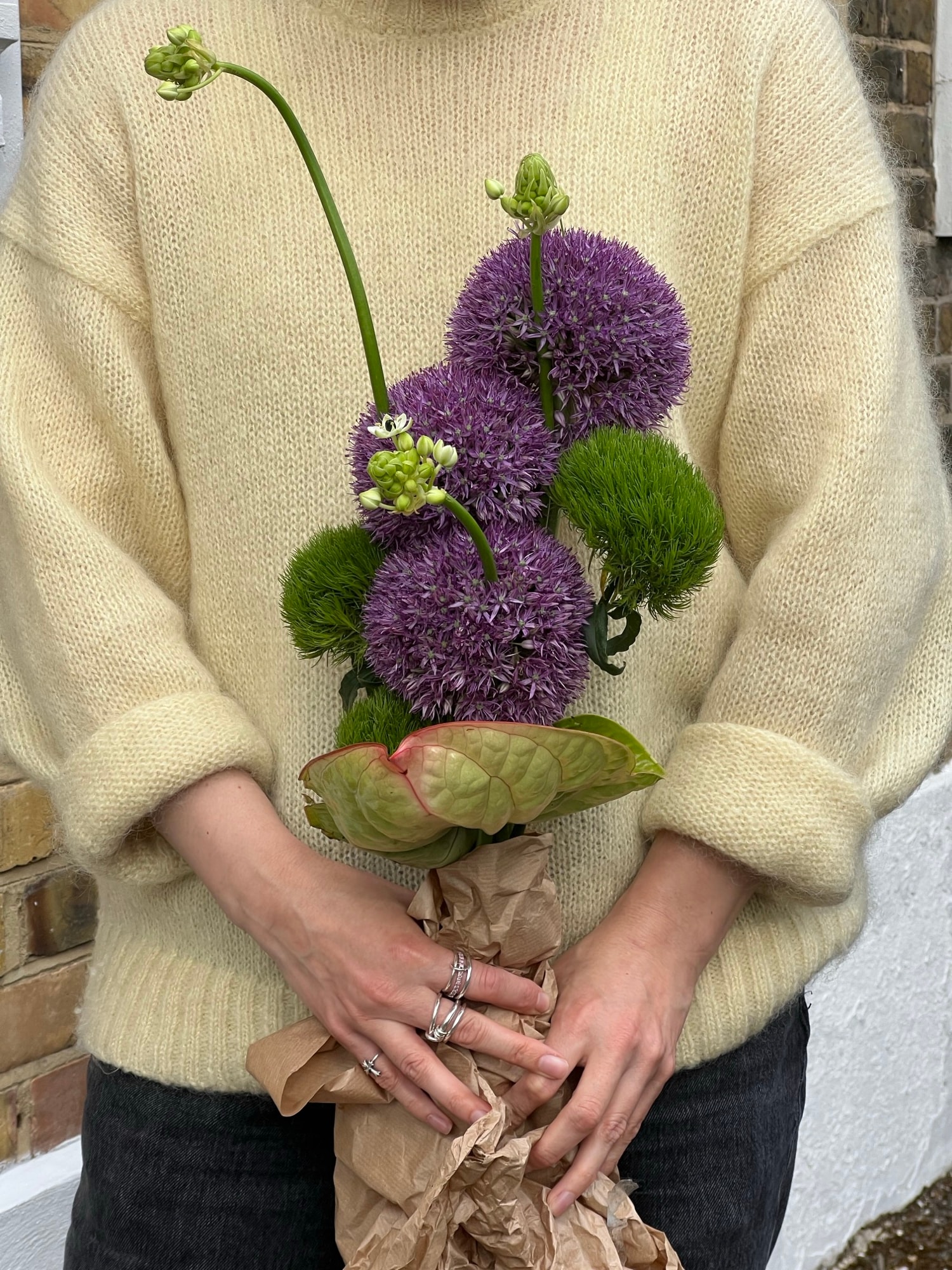 Hands in a cream fuzzy sweater holding a kraft-wrapped bouquet of purple alliums and green blooms