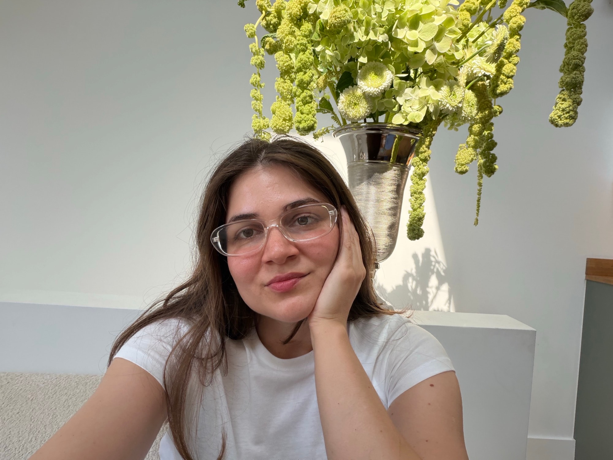Founder in glasses and white tee posing beside tall green flowers in a silver vase