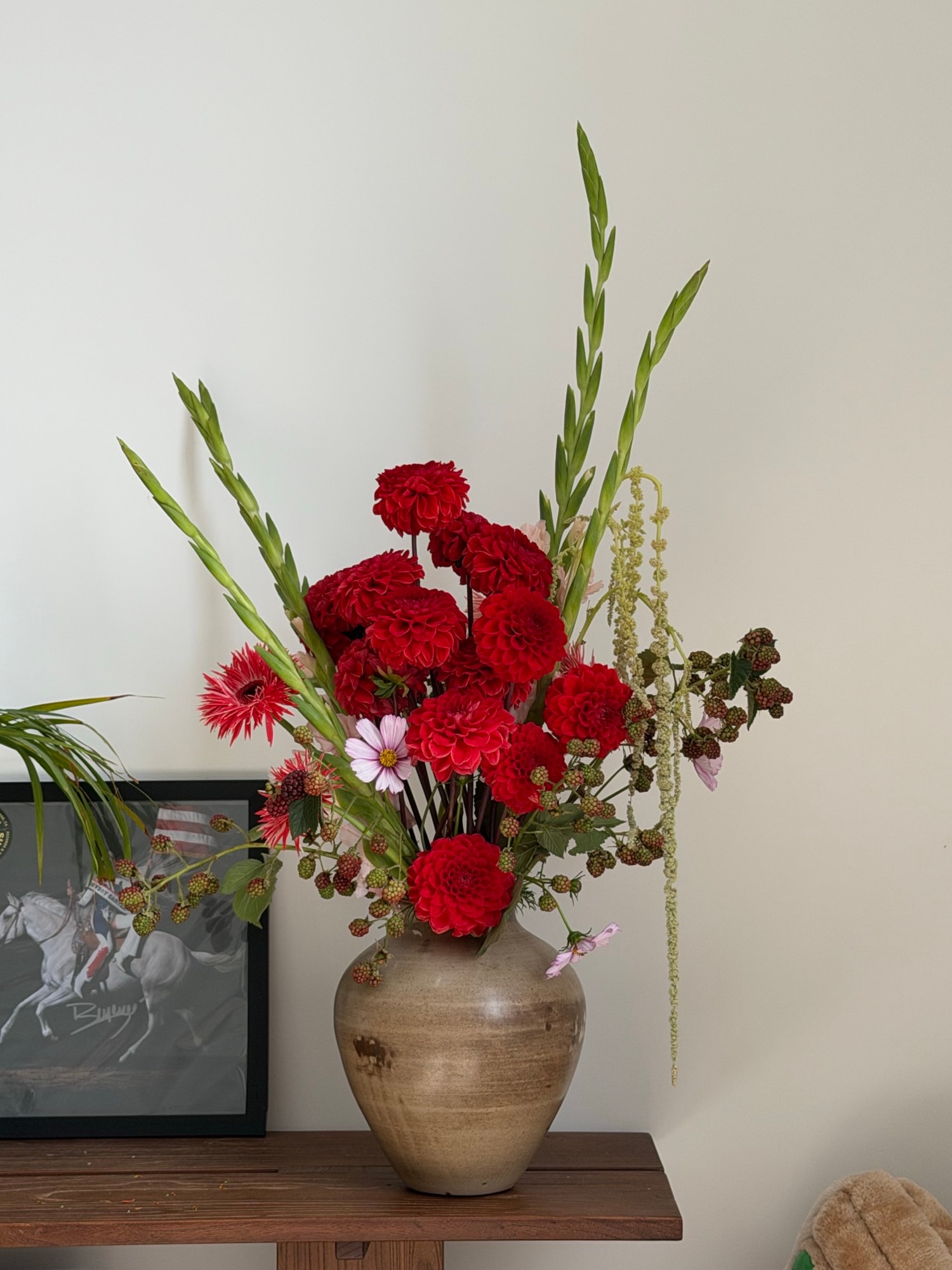 Asymmetric arrangement of red dahlias, cosmos, and green gladiolus in a beige ceramic vase on dark wood