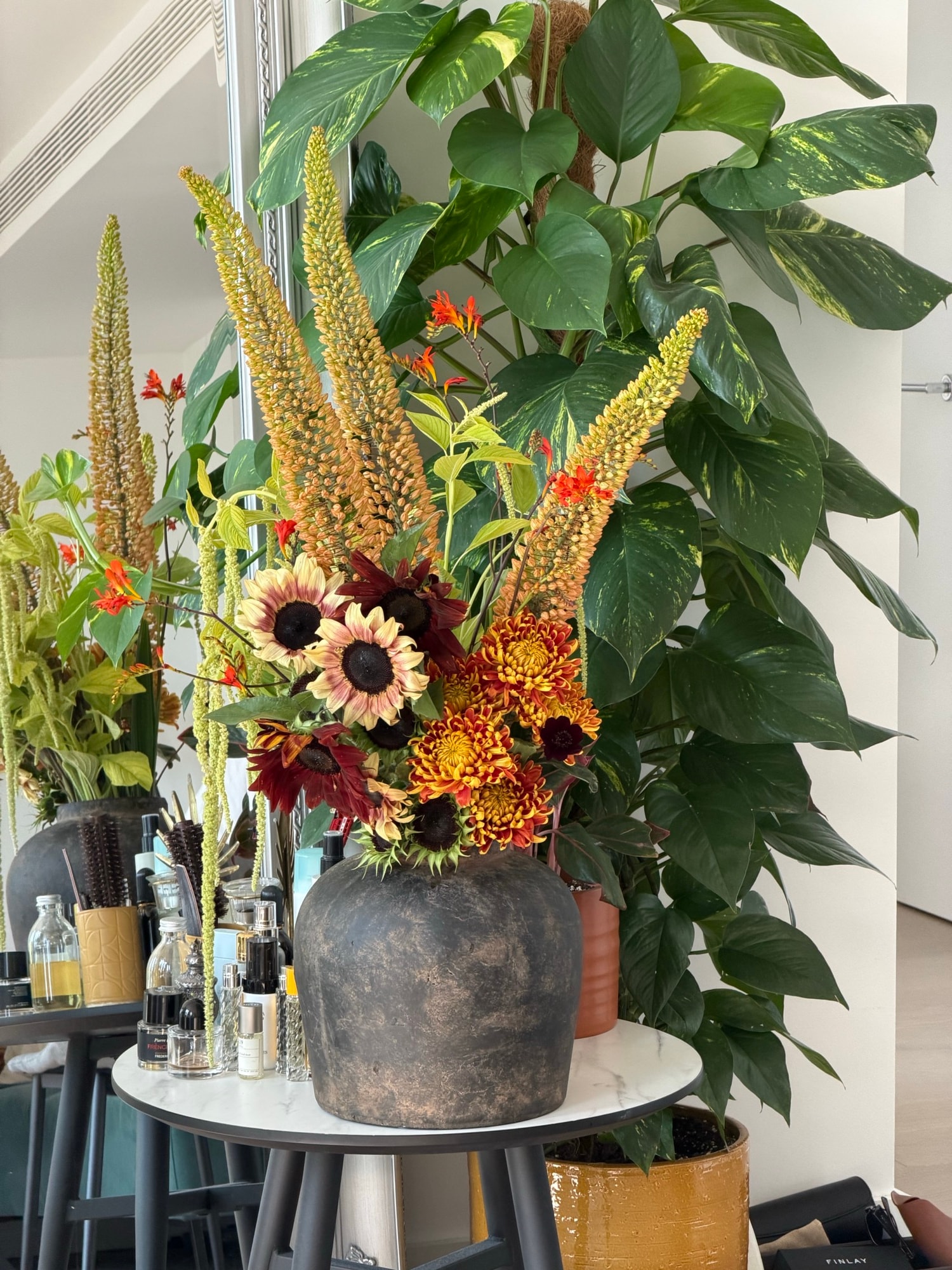 Sunflowers and foxtail lilies in a charcoal vase on a white table with leafy plant and studio supplies behind