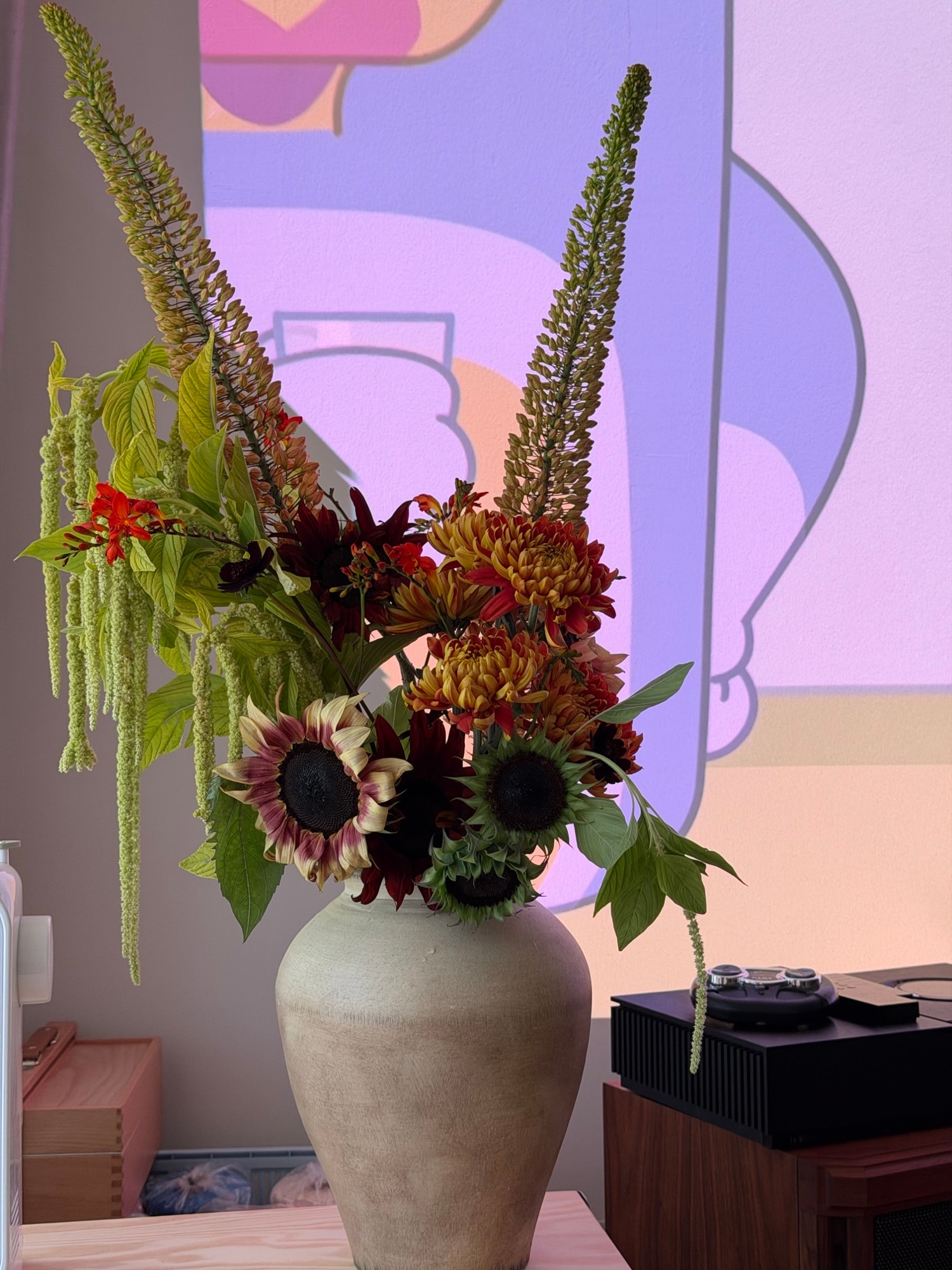 Tall off-white vase with sunflowers, bronze chrysanthemums and green spikes beside a projected landscape