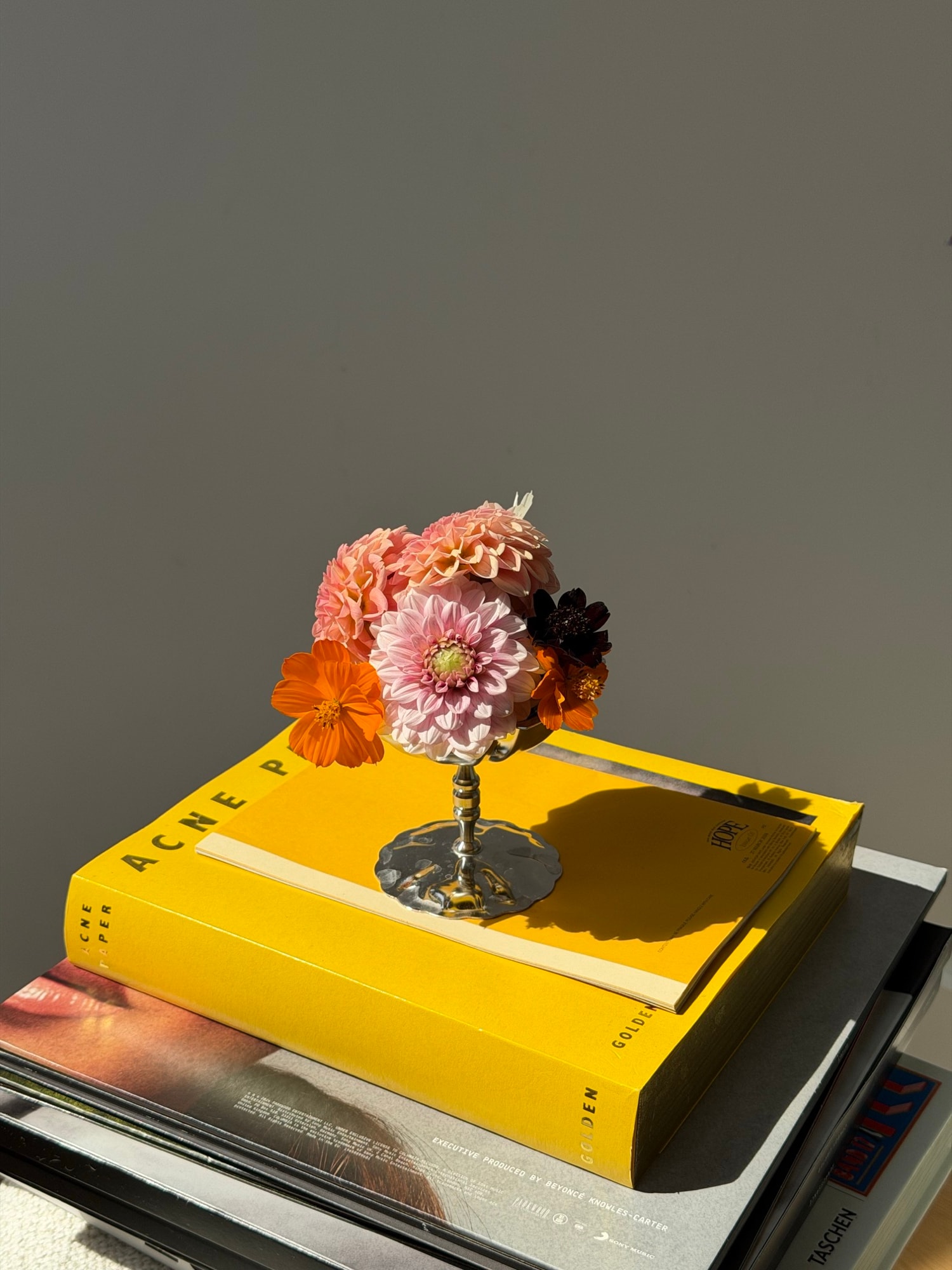 Pink dahlias and orange blooms in silver chalice on yellow Acne Paper book