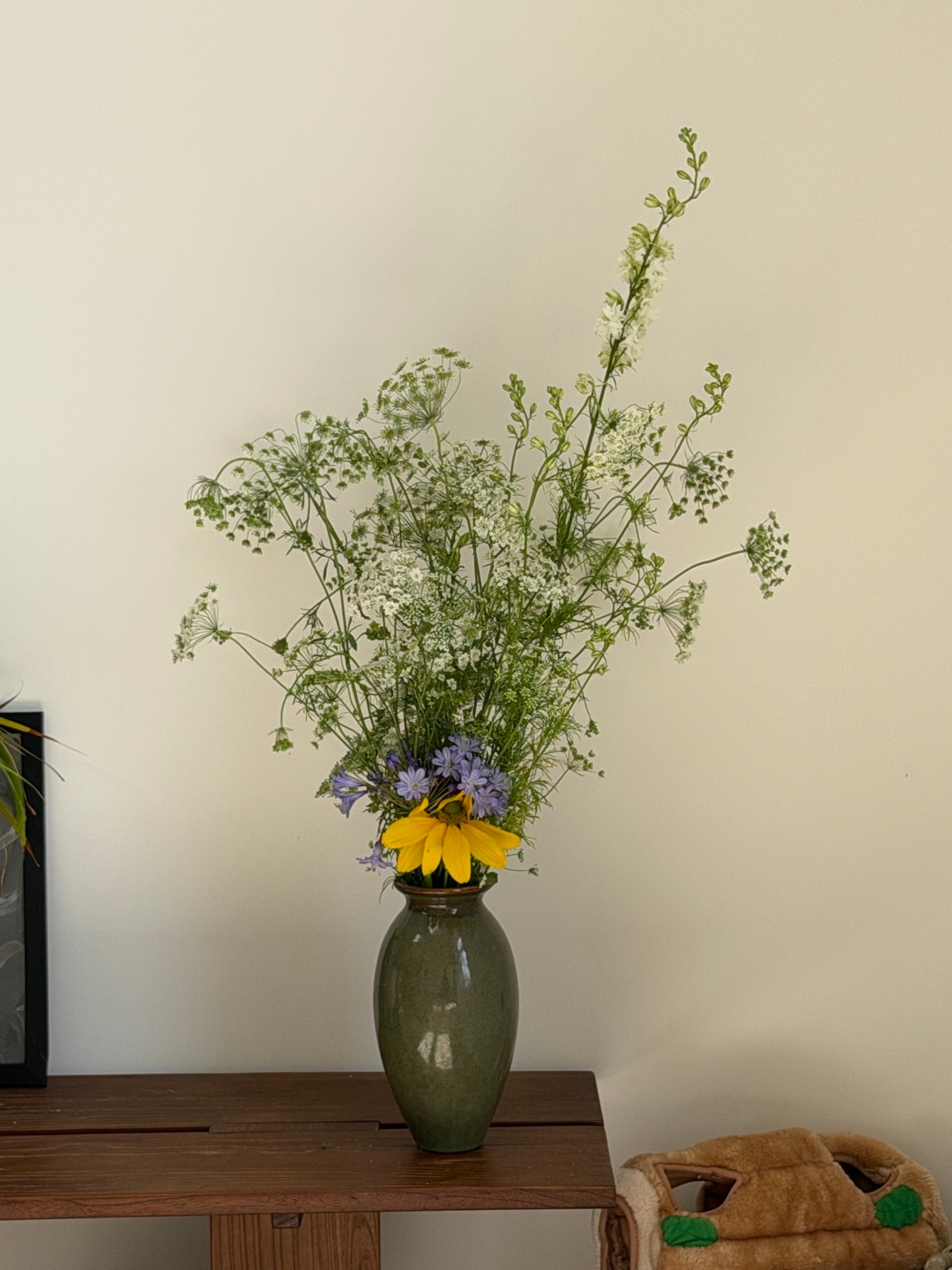 Wild white umbels and yellow bloom in olive vase on wood shelf